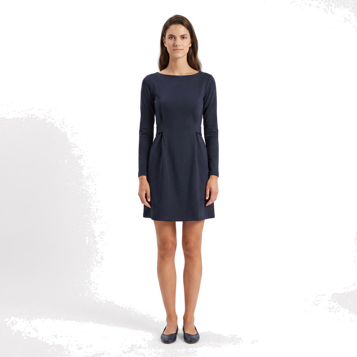 A woman modeling a navy blue long-sleeve shift dress with a boat neckline and flared skirt, paired with navy flats, against a white studio background.