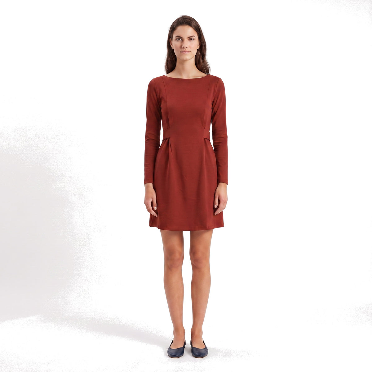 A woman modeling a rust-red long-sleeve shift dress with a boat neckline and flared skirt, paired with dark navy flats, against a white studio background.