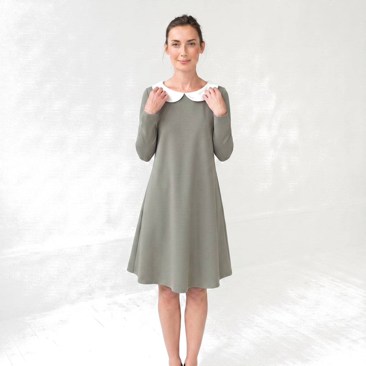 A woman modeling a long-sleeve knee-length dress in muted sage green with a contrasting white Peter Pan collar, standing against a light gray textured studio backdrop.