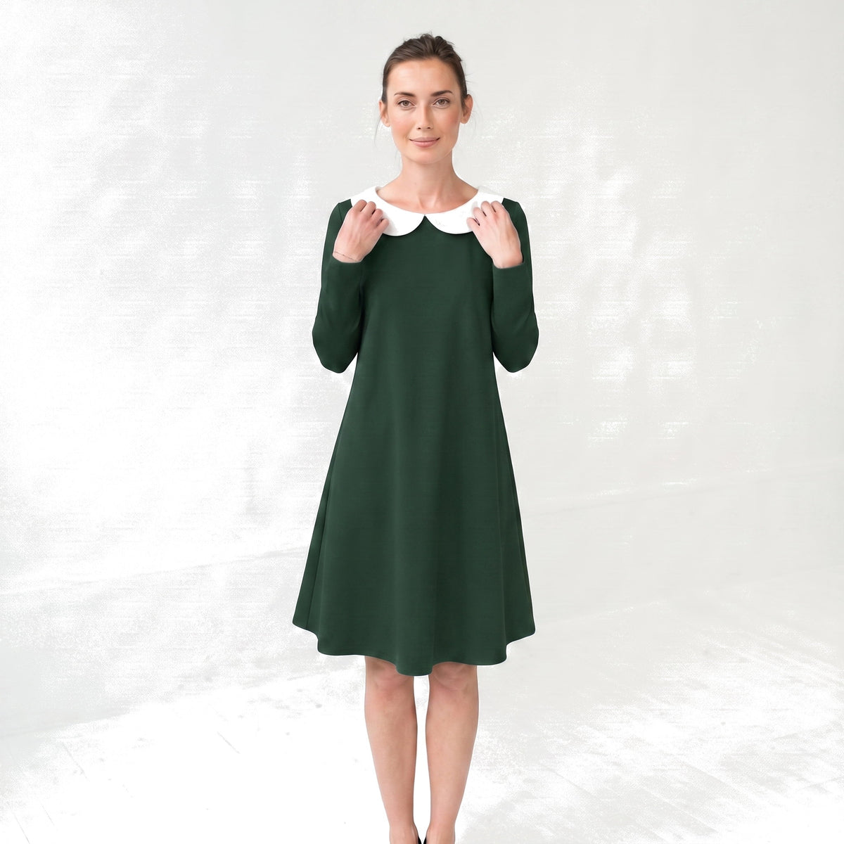 A woman modeling a dark green long-sleeve A-line dress with a contrasting white Peter Pan collar, standing against a light gray textured studio backdrop.