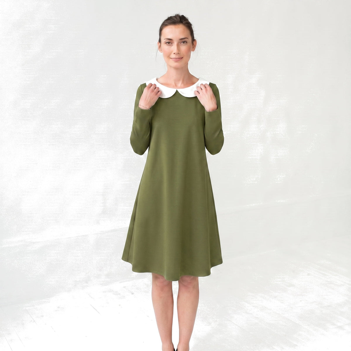 A woman modeling a long-sleeve olive green dress with a contrasting white Peter Pan collar, standing against a light gray textured studio backdrop.