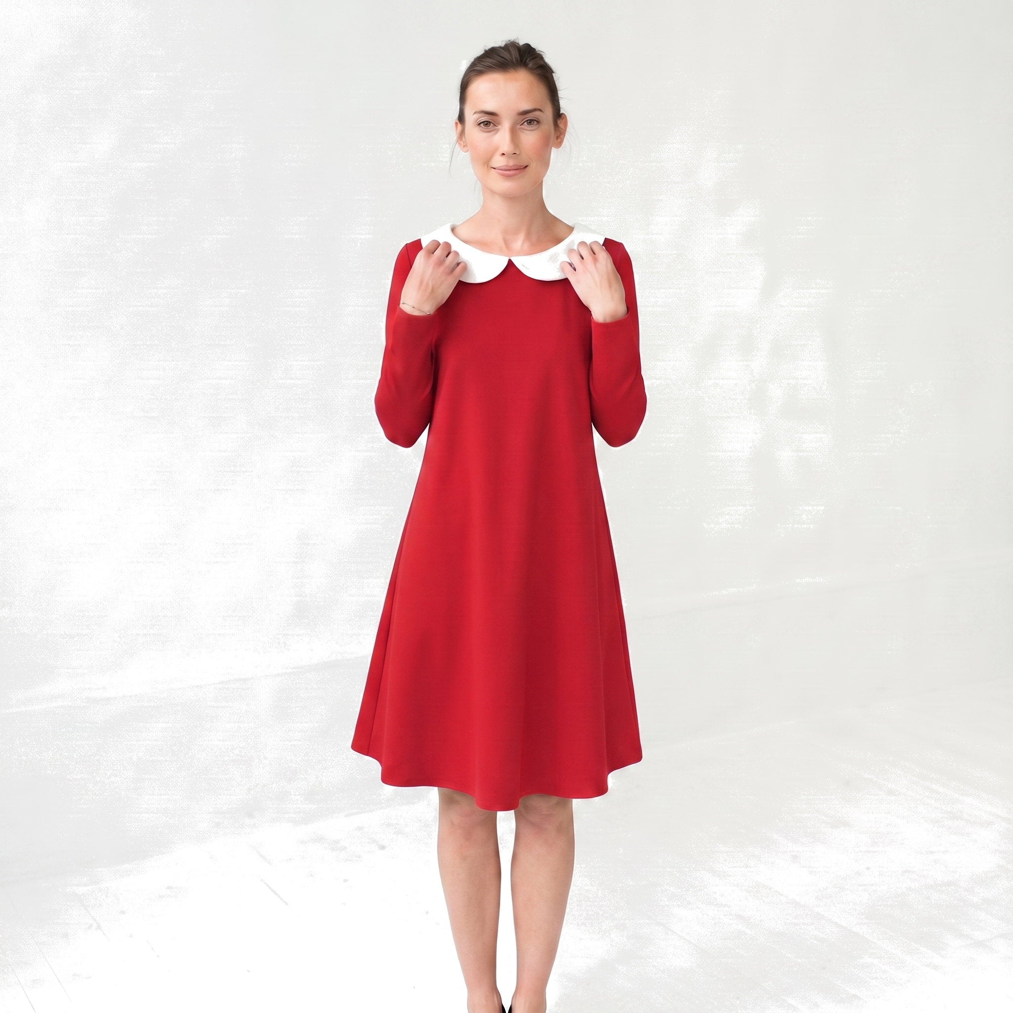 A woman modeling a red long-sleeve A-line dress with a contrasting white Peter Pan collar, standing against a light gray textured studio backdrop.