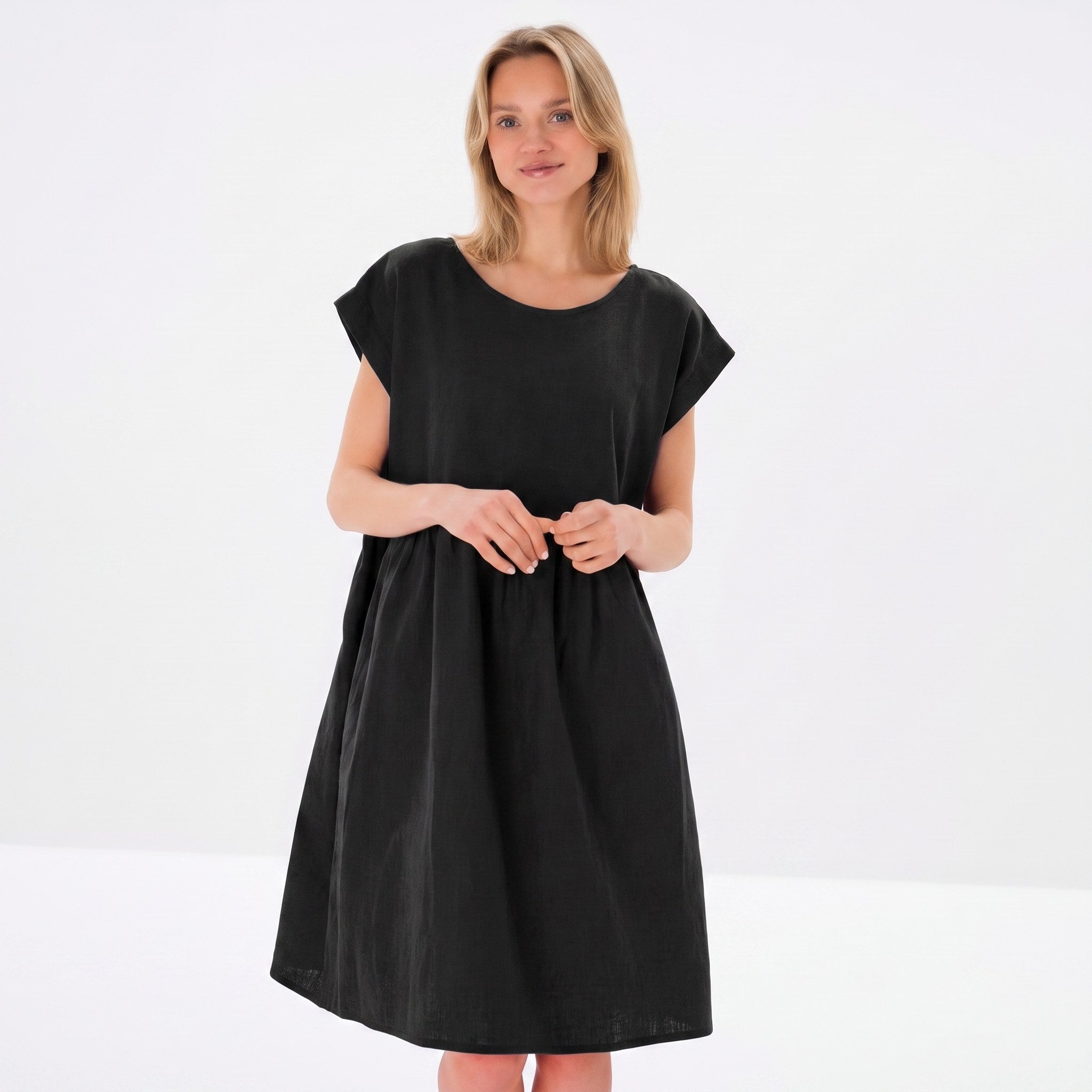 A woman modeling a loose-fitting black dress with short cap sleeves and a round neckline, photographed against a clean white studio background.