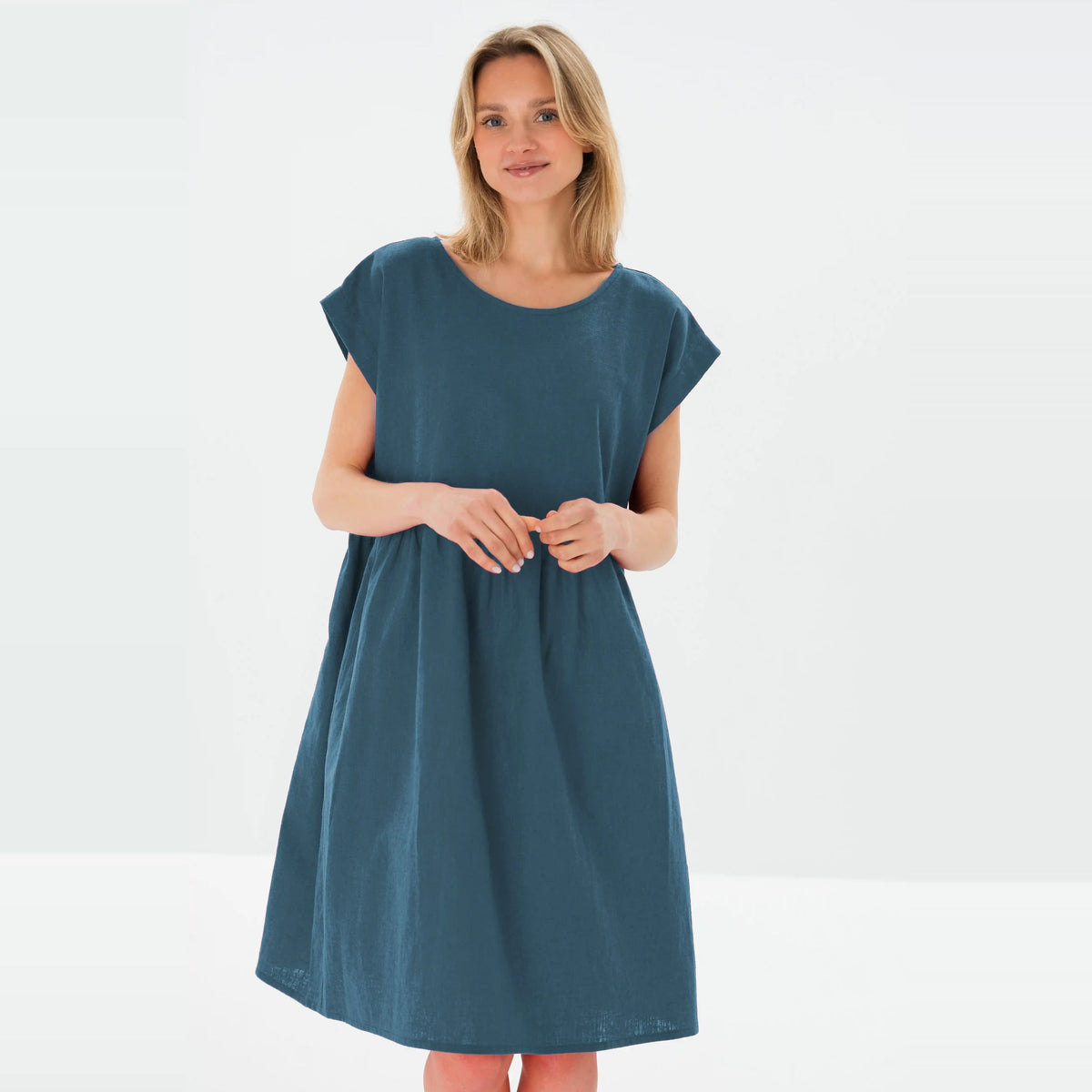 A woman modeling a loose-fitting, short-sleeve A-line dress in a muted teal blue color, standing against a clean white studio background.