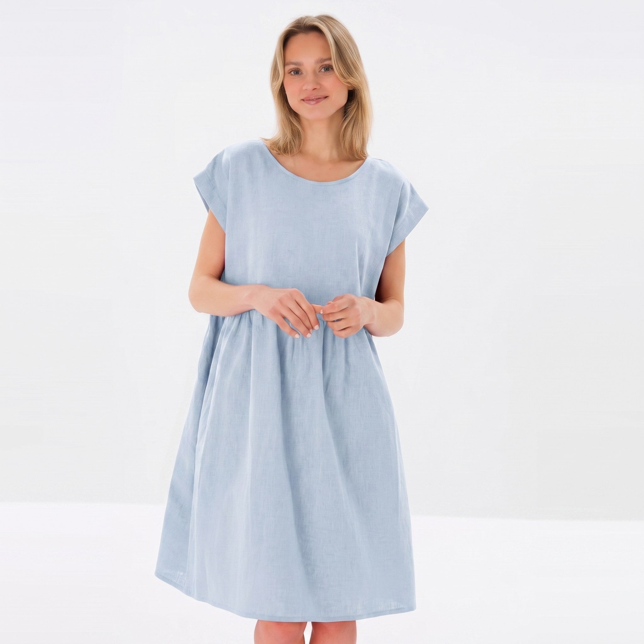 A woman modeling a light blue linen dress with short cap sleeves and a relaxed, gathered silhouette against a plain white studio background.
