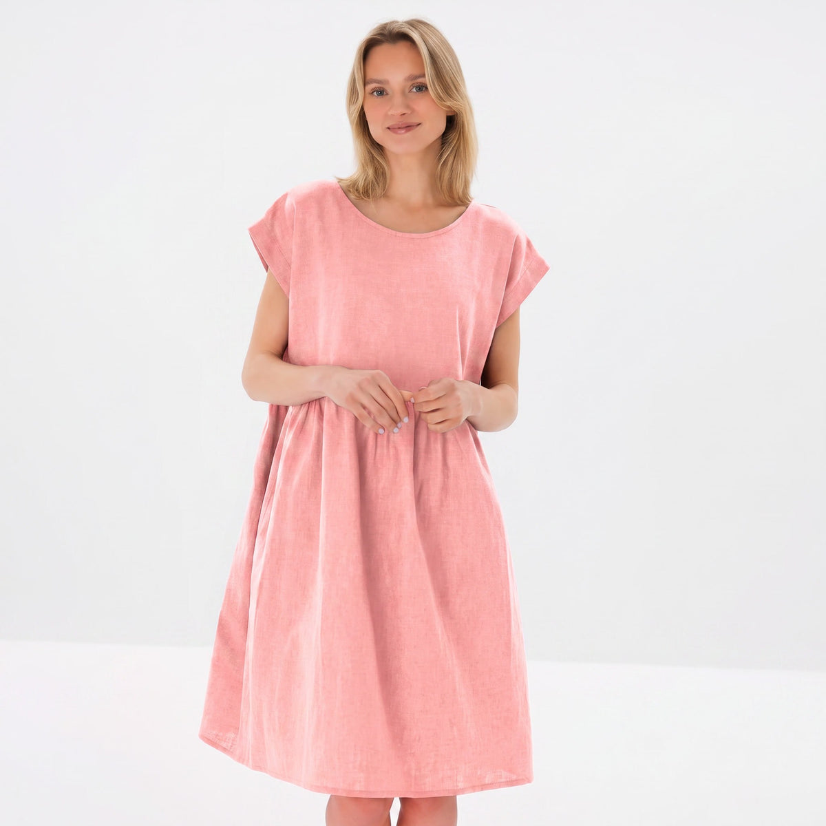 A woman modeling a light pink linen dress with short cap sleeves and a relaxed, gathered silhouette against a plain white studio background.