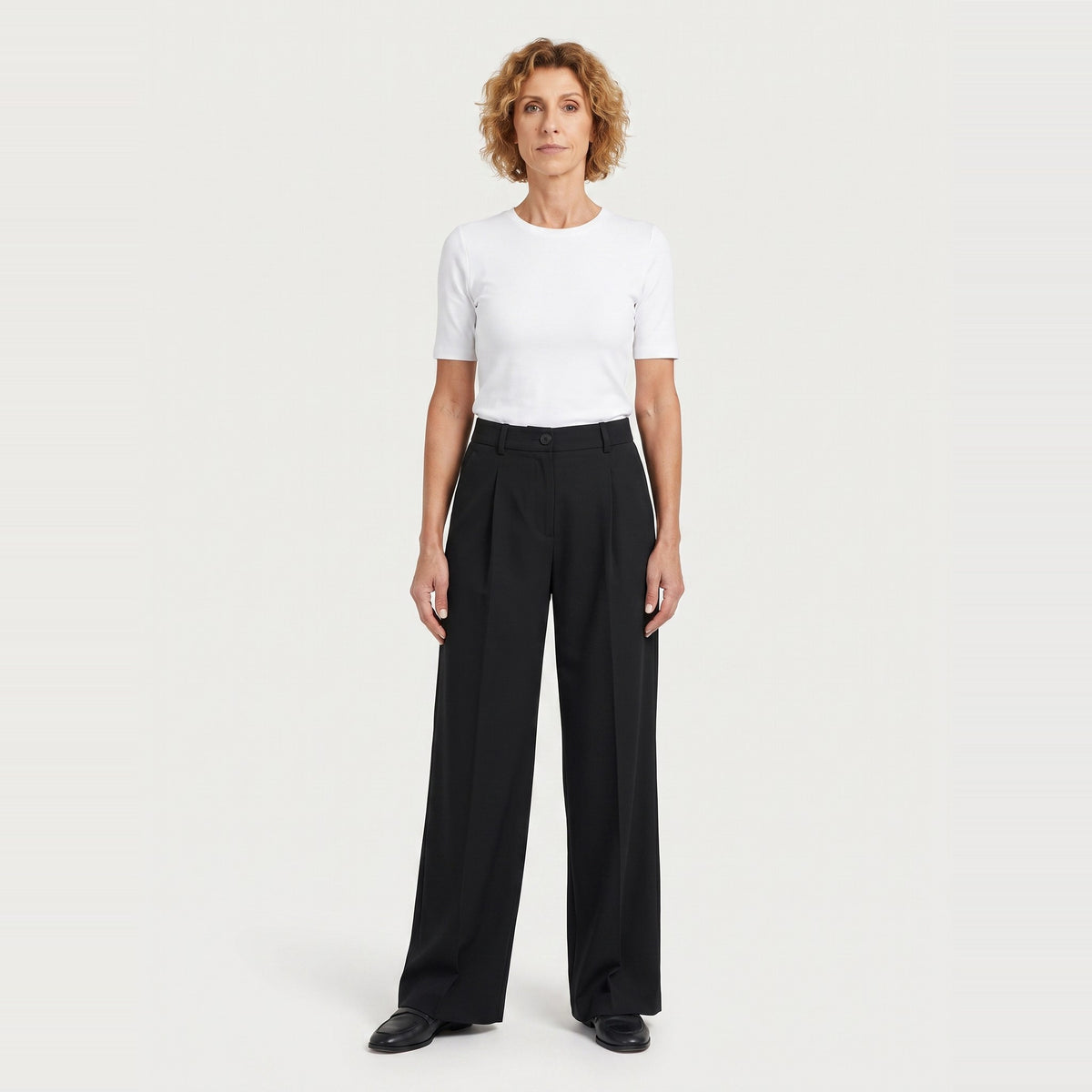 A woman stands against a light gray studio background wearing a white short-sleeve crewneck T-shirt and black wide-leg trousers.