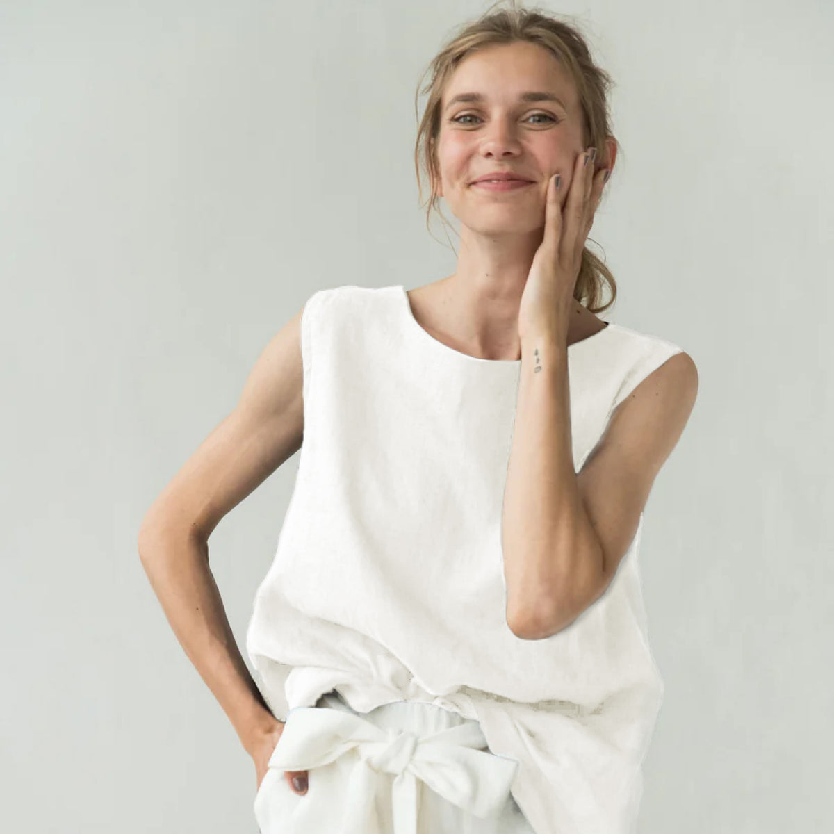 A woman wearing a sleeveless white linen top and matching white bottoms tied at the waist, posing against a light gray background.