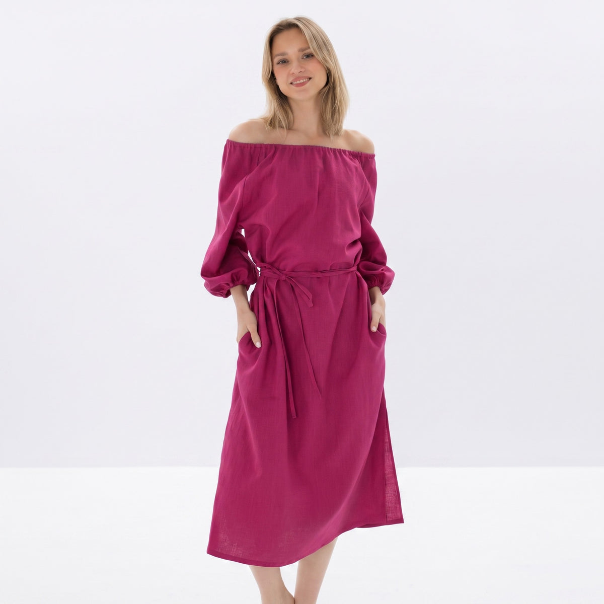 A woman modeling an off-the-shoulder midi dress in a rich magenta-pink color, featuring long sleeves, a matching fabric tie belt at the waist, and side pockets, set against a clean white studio background.