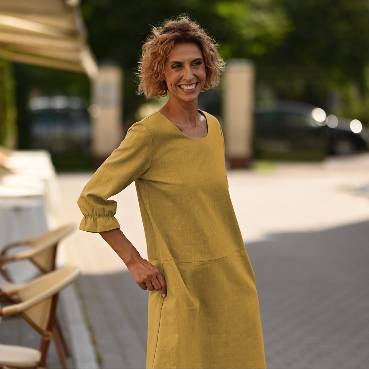 A woman wearing a mustard yellow linen dress with three-quarter sleeves and a round neckline, standing outdoors on a paved path with cafe chairs and trees in the background.