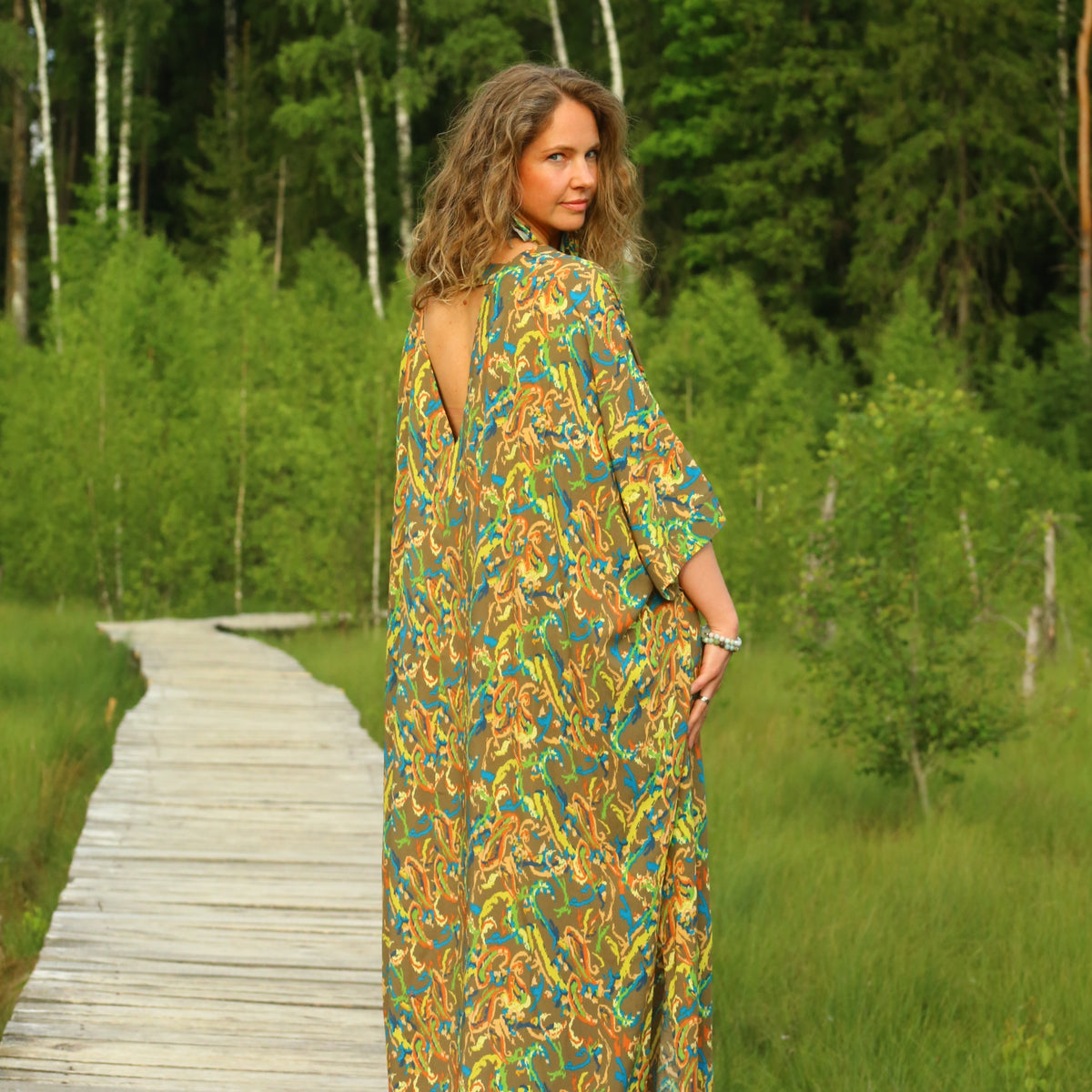 A woman stands on a wooden boardwalk in a lush green forest, wearing a long-sleeved maxi dress with a colorful abstract pattern in yellow, green, and blue.