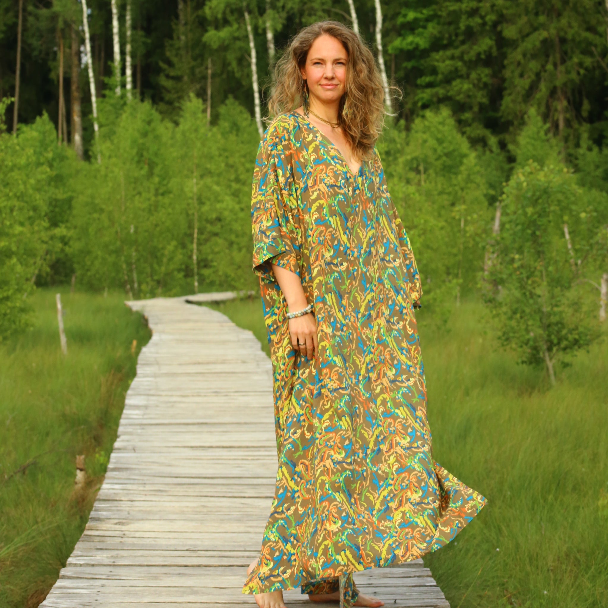 A woman stands on a wooden boardwalk in a lush green forest, wearing a long, loose kaftan-style dress with a colorful abstract leaf pattern in yellow, orange, blue, and green.