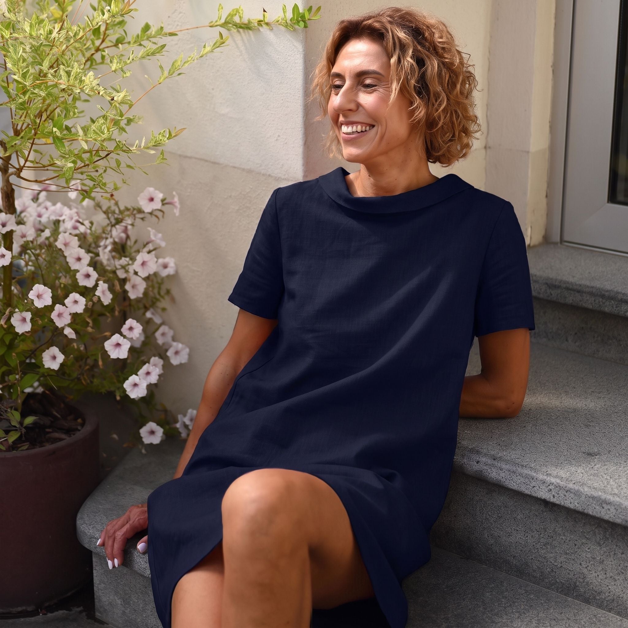 A woman with curly light brown hair sits smiling on outdoor stone steps, wearing a loose short-sleeve navy dress. A potted flowering plant and a light-colored stucco wall are visible in the background.