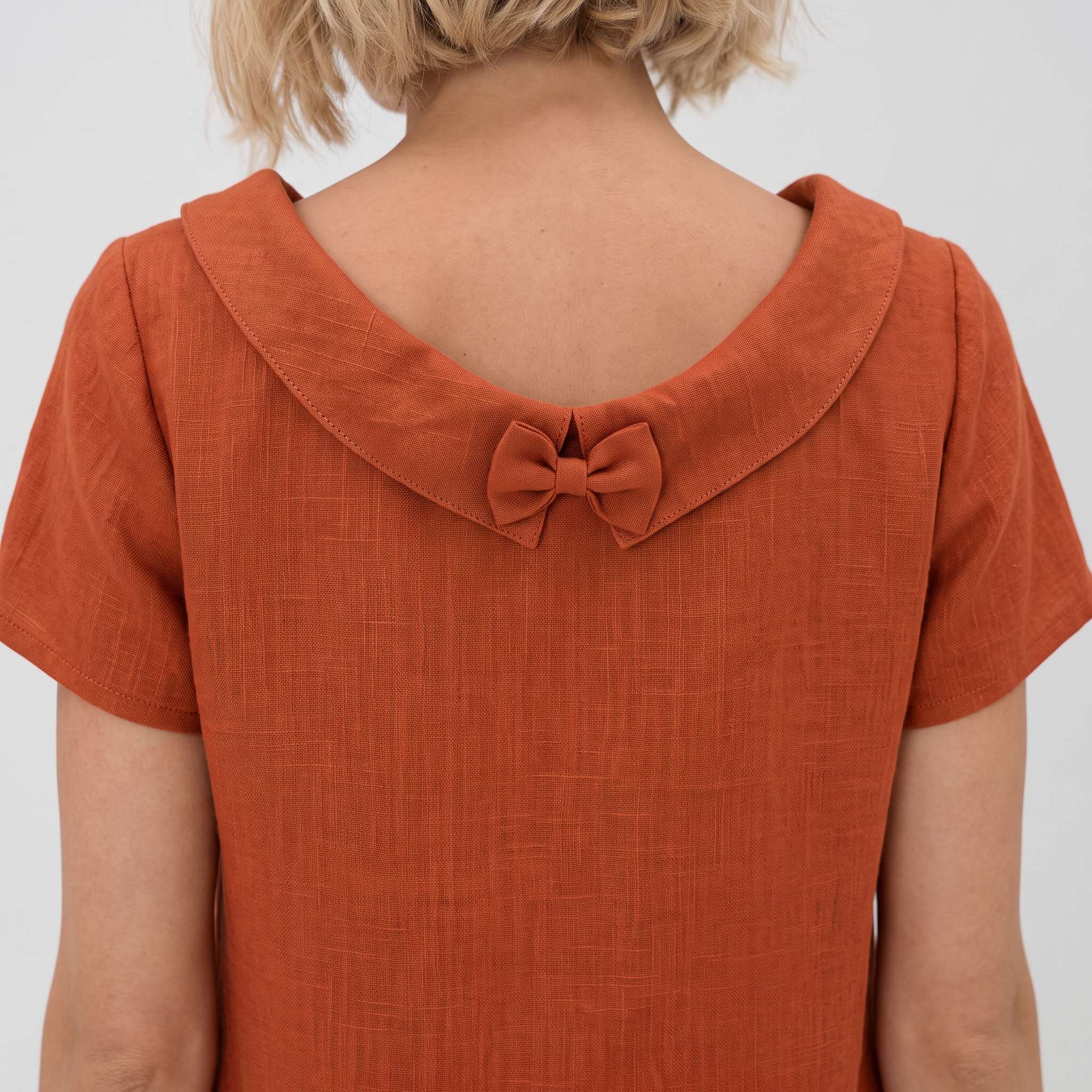 A close-up lifestyle shot of a woman with curly light brown hair wearing an orange short-sleeve dress with a wide collar and bow detail on the back.