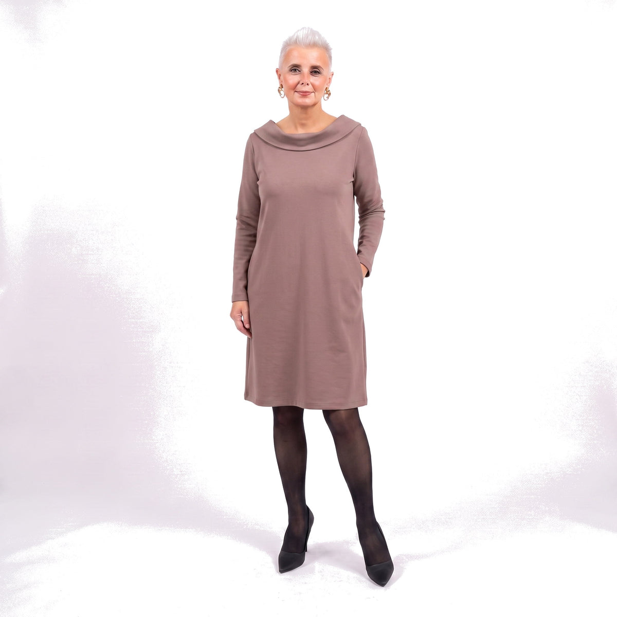 A woman modeling a long-sleeve, knee-length cowl-neck dress in a muted taupe-brown color, paired with black tights and black pointed-toe heels, against a white studio background.
