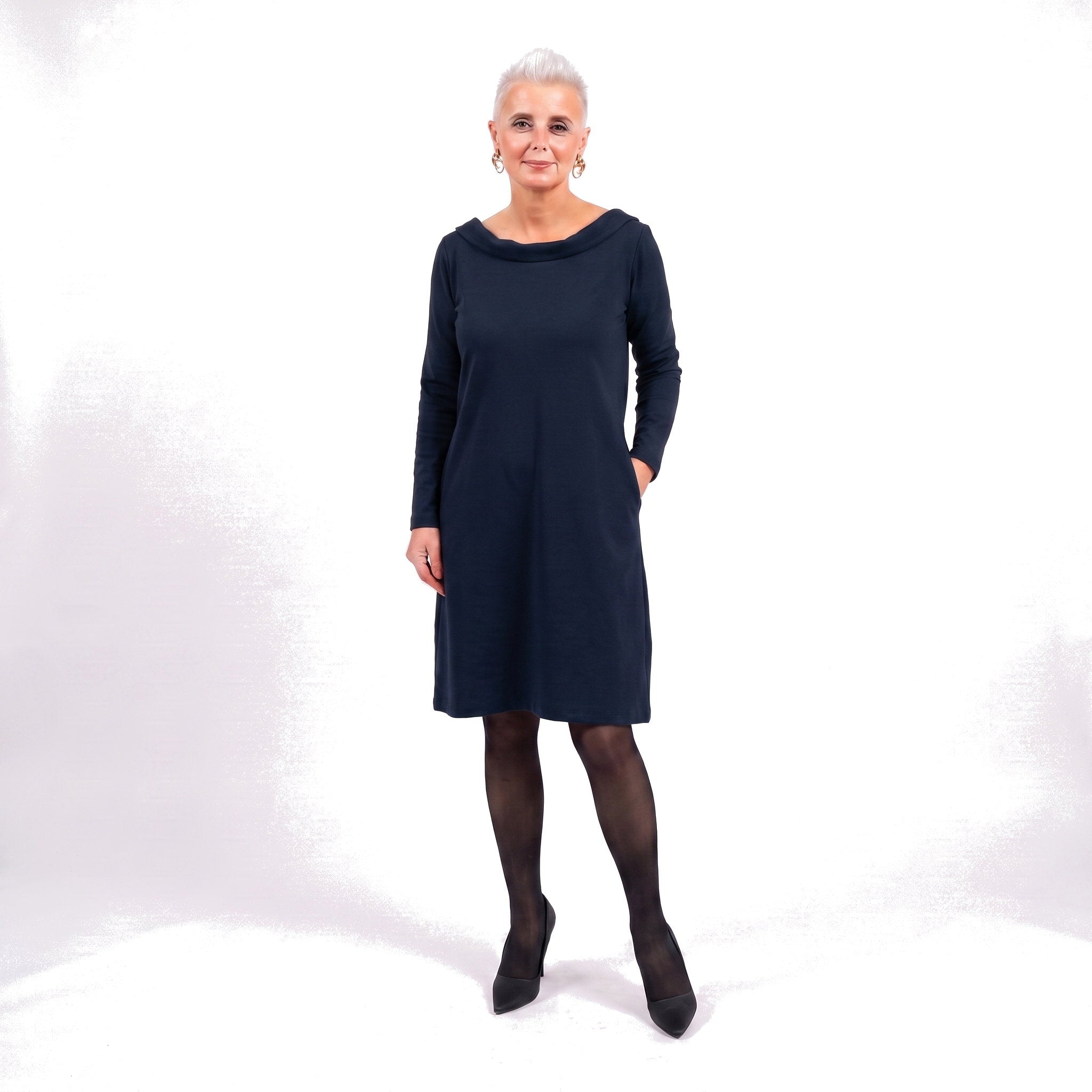 An older woman with short gray hair models a dark navy blue long-sleeve shift dress with pockets, paired with black tights and black pointed-toe heels, against a white studio background.