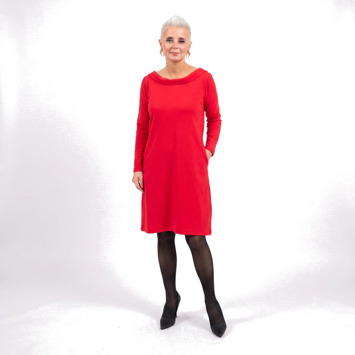 A woman modeling a bright red long-sleeve shift dress with black tights and black pointed shoes, standing against a white studio background.