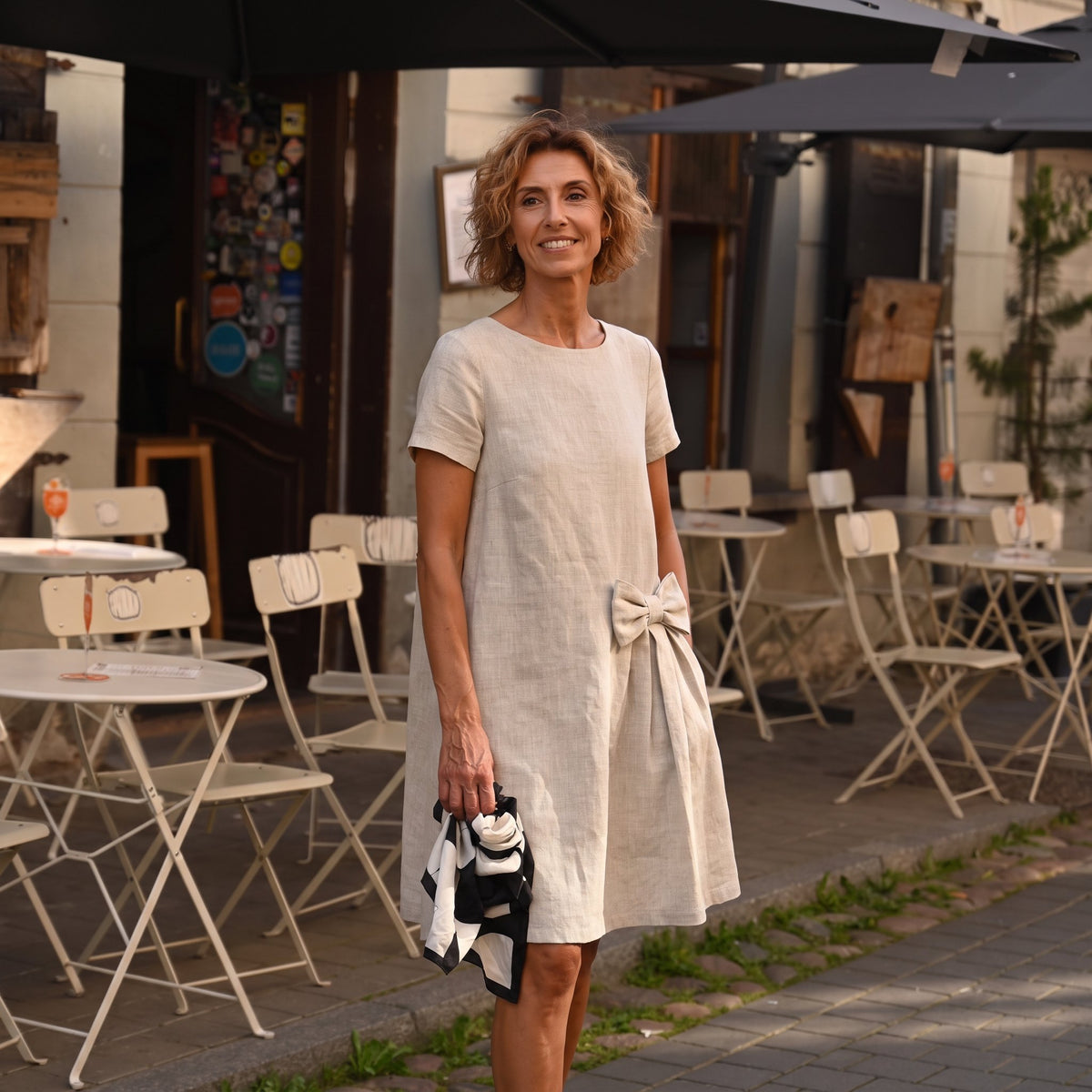 A woman stands outdoors at a cafe wearing a short-sleeved beige linen dress with a large bow detail at the waist. She is holding a black and white patterned handbag, with cafe tables and chairs visible in the background.