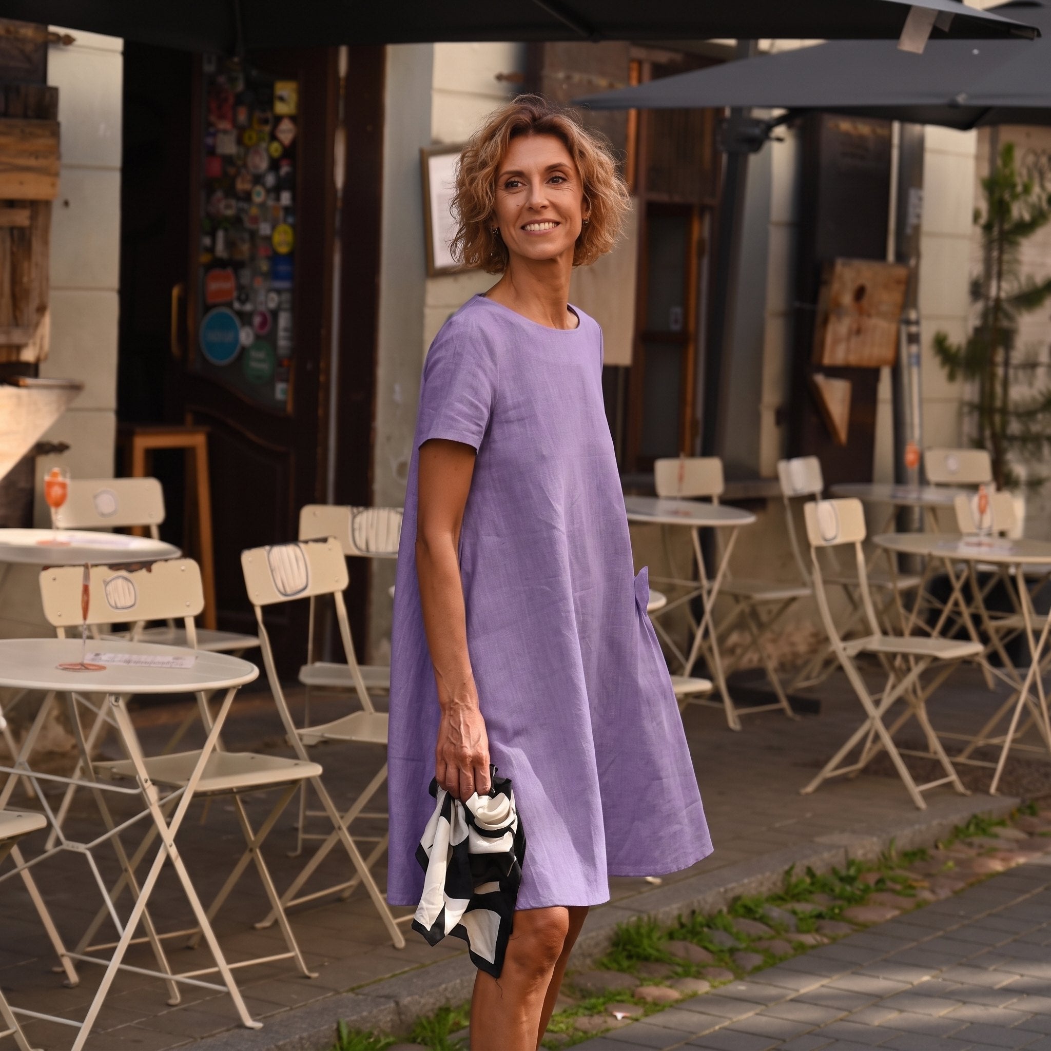 A woman wearing a lavender short-sleeve linen dress stands on an outdoor cafe patio, holding a black and white patterned clutch bag.