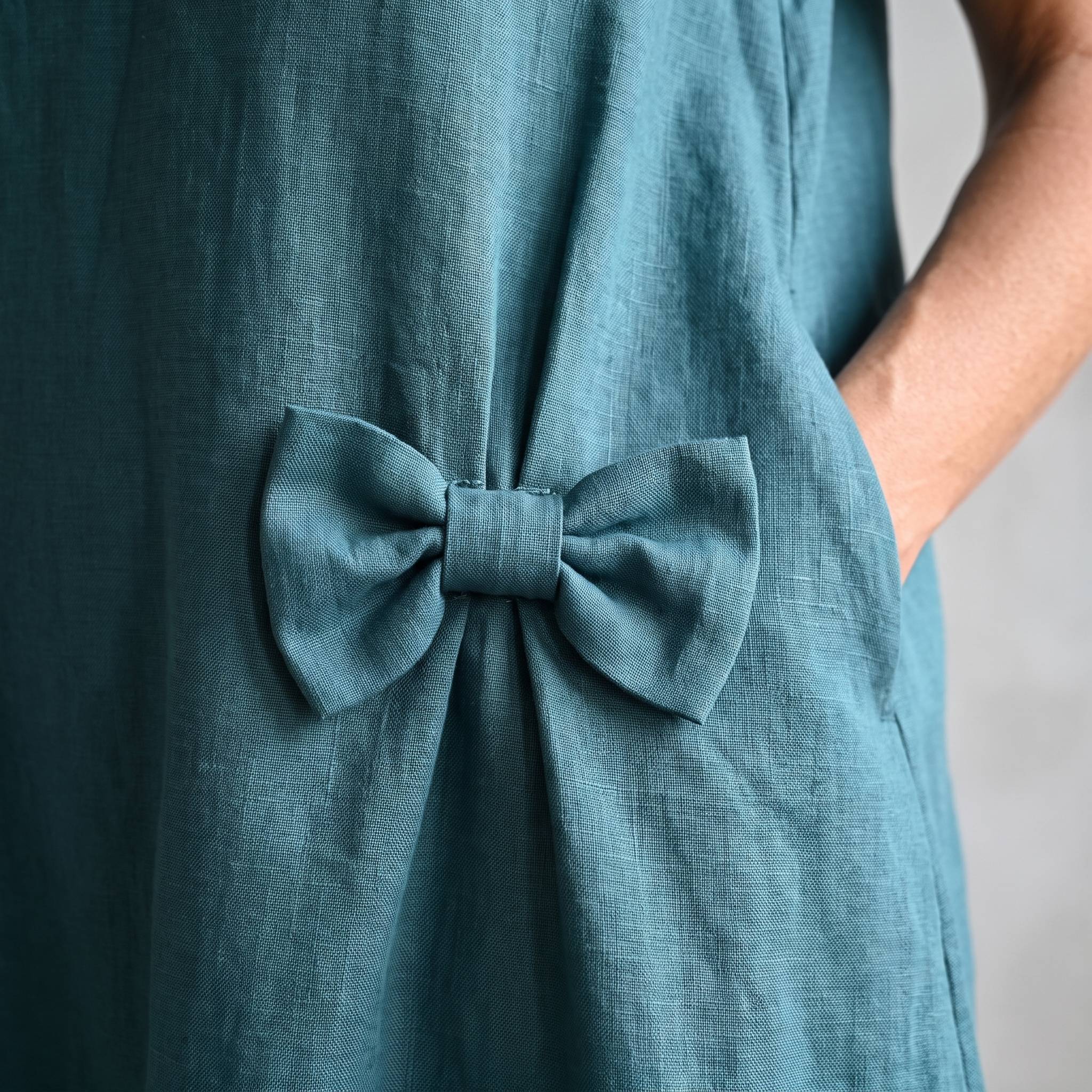 A close-up of a teal blue linen dress featuring a large fabric bow detail at the center back, worn by a model whose arm is visible on the right side.