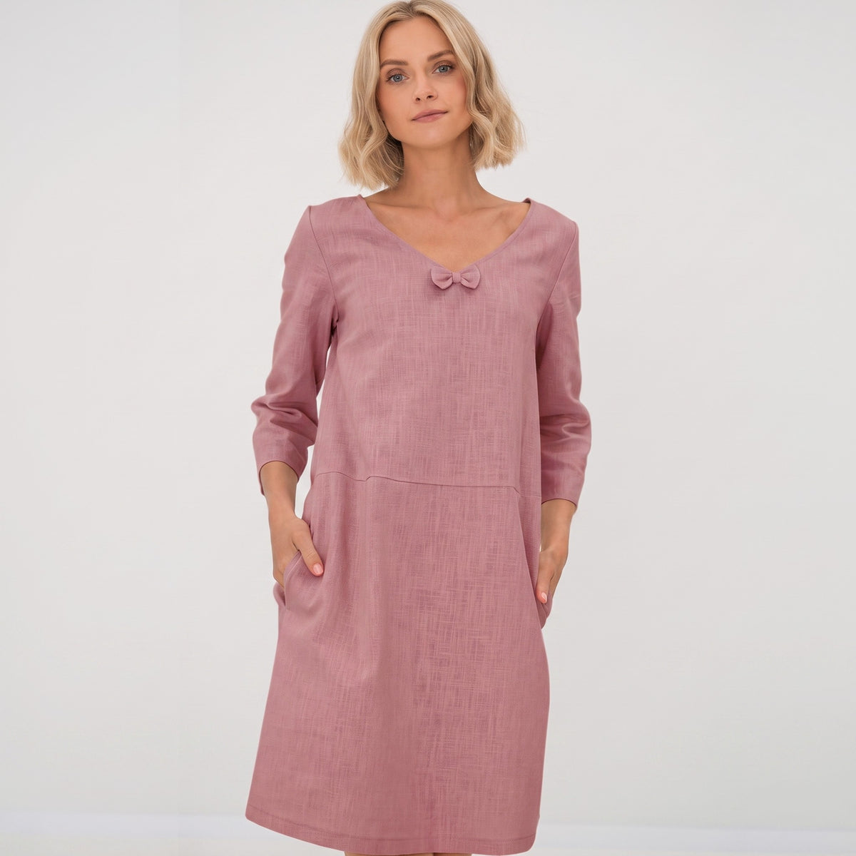 A woman modeling a dusty rose pink long-sleeve linen dress with a small bow detail at the neckline, set against a plain light gray studio background.