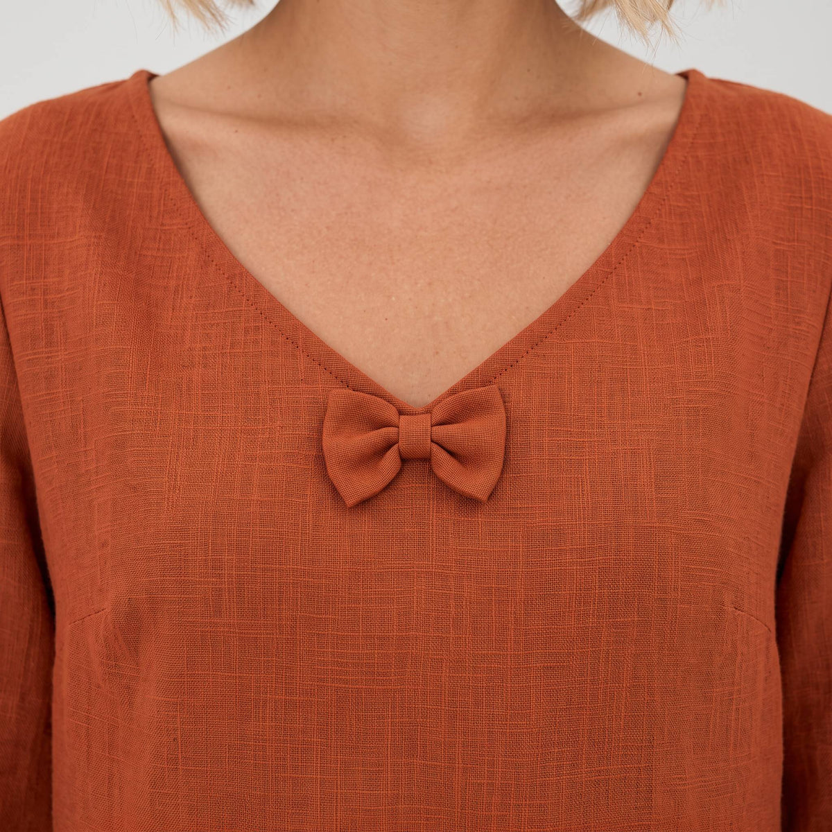 Close-up of a terracotta orange linen top with a V-neck and a matching fabric bow detail at the center of the neckline, worn by a model.