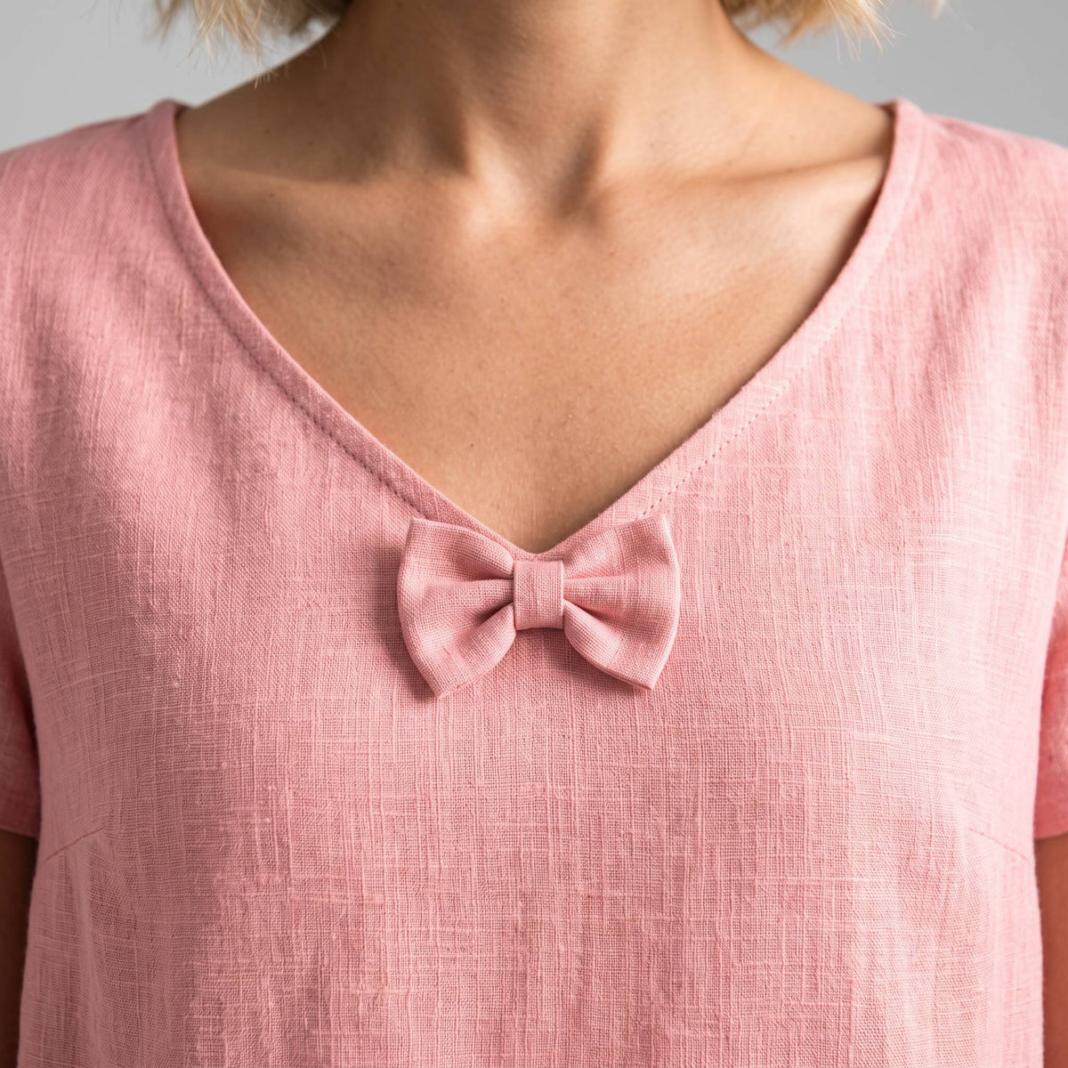 A close-up of a person wearing a light pink linen V-neck top with a matching fabric bow detail at the center of the neckline.