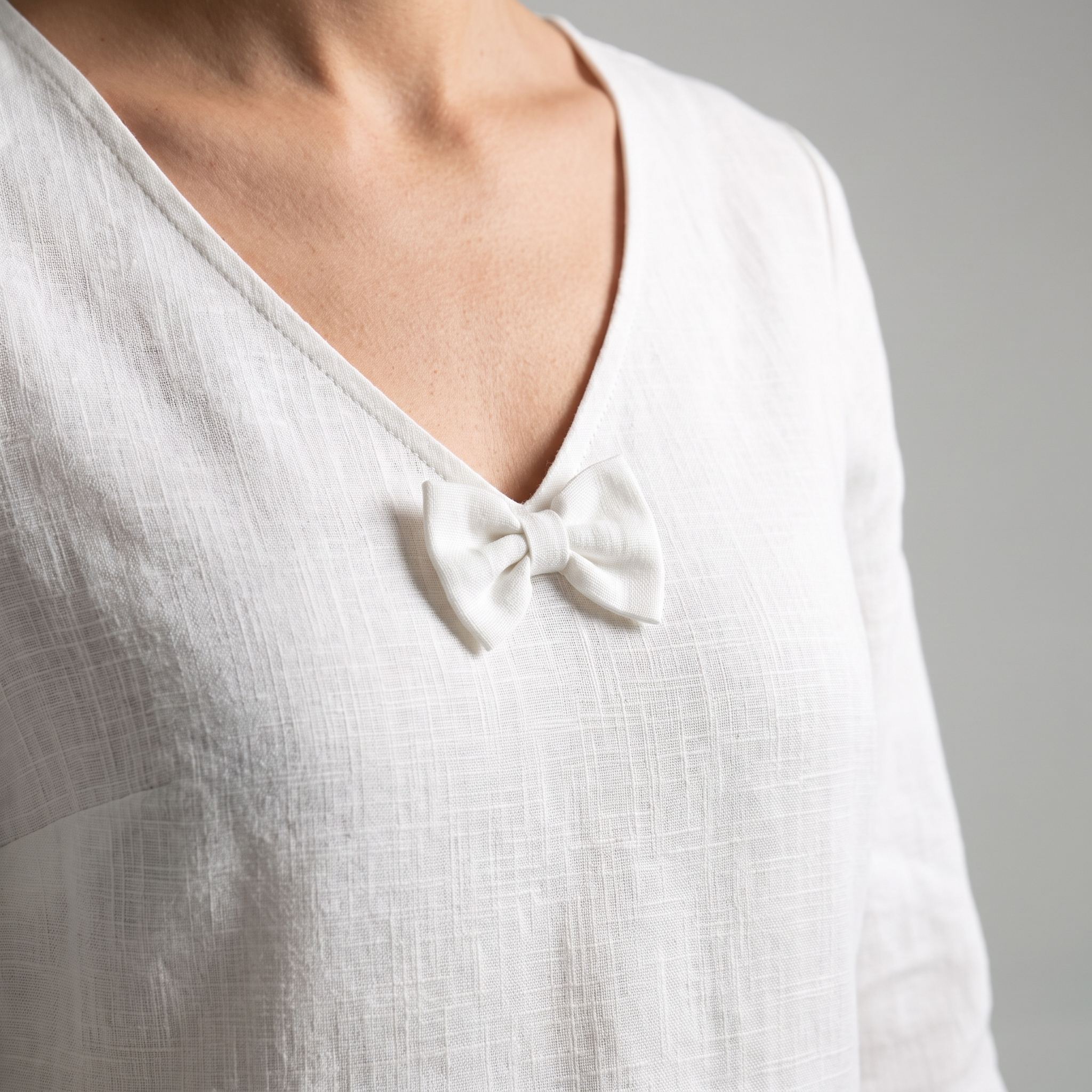 Close-up of a person wearing a white textured linen top with a small bow detail at the V-neck, against a neutral gray background.