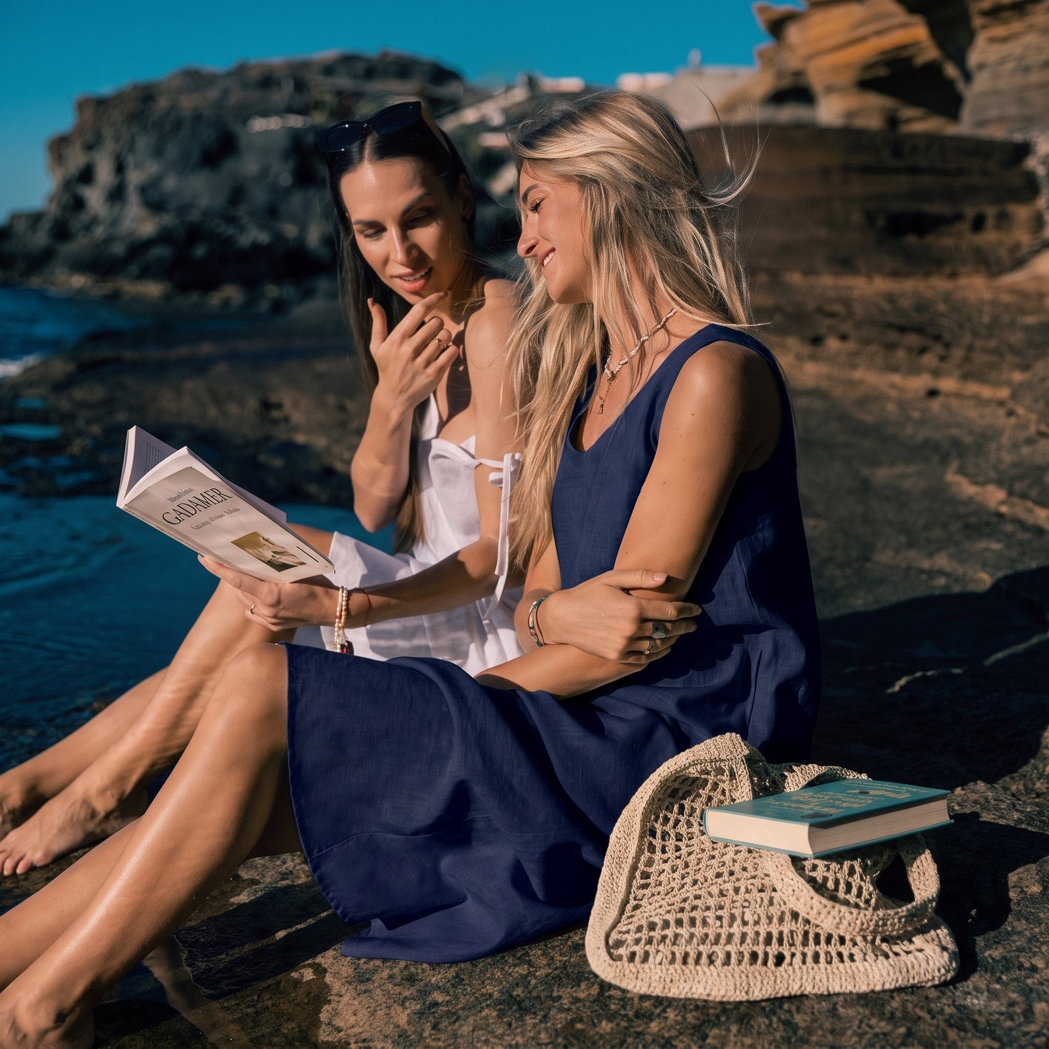 Two women sitting on a rocky beach reading a book, with one wearing a navy blue sleeveless dress and the other in a white dress. A woven straw bag with a book rests beside them, and the ocean and cliffs are in the background.