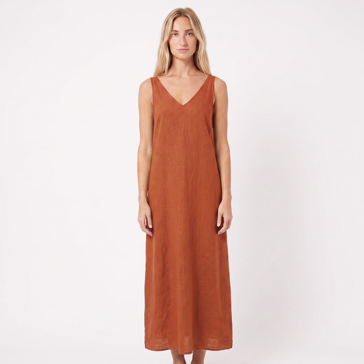 A woman modeling a sleeveless, V-neck maxi dress in a warm rust orange color, standing against a plain white studio background.