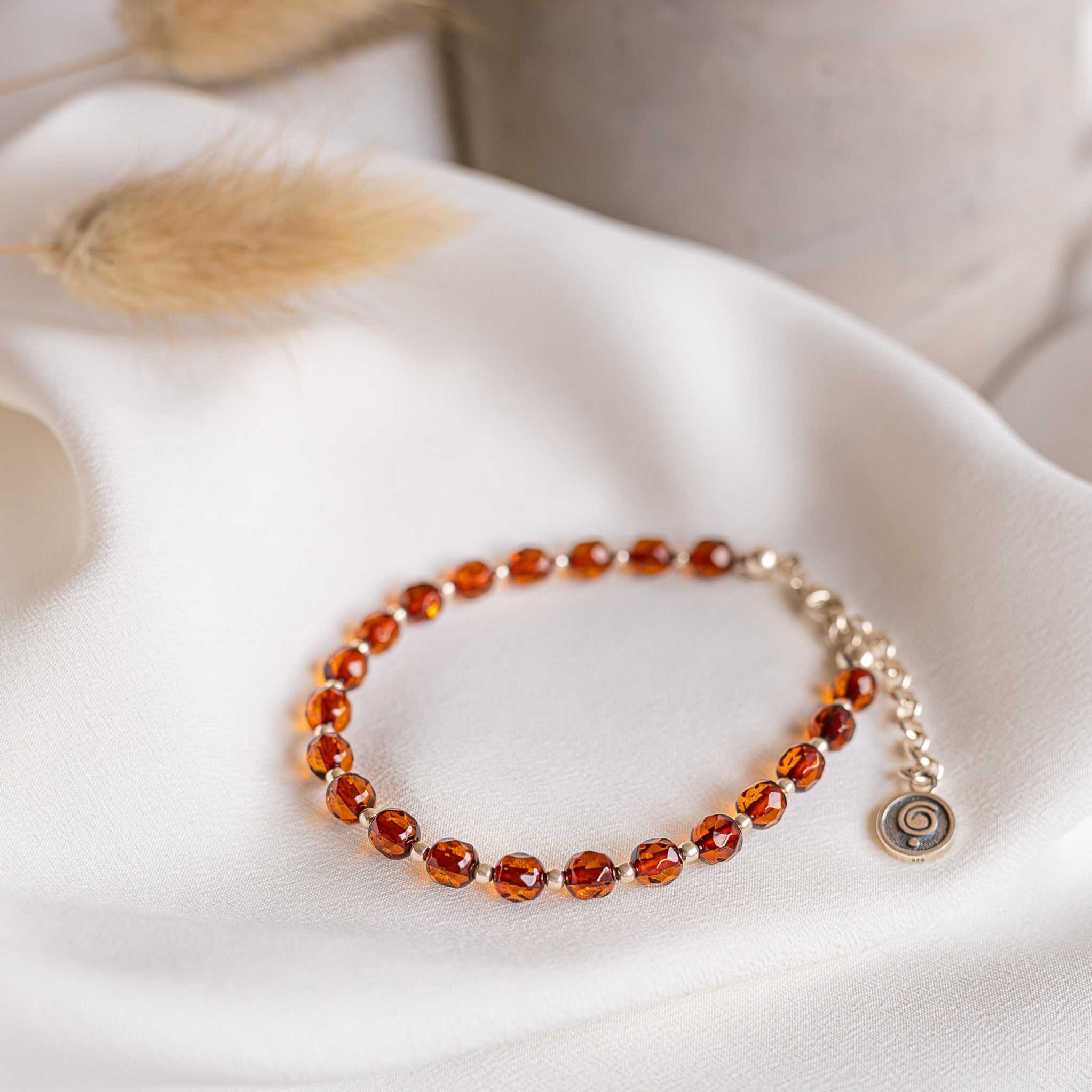 A beaded bracelet with faceted amber-orange beads and a small round charm, displayed on draped white fabric with soft decorative dried grass in the background.