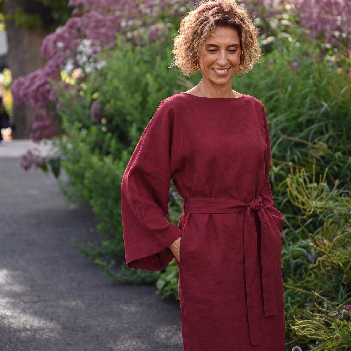A woman wearing a loose-fitting, long-sleeved burgundy dress with a matching waist tie, standing outdoors in a garden setting with green foliage and purple flowers.